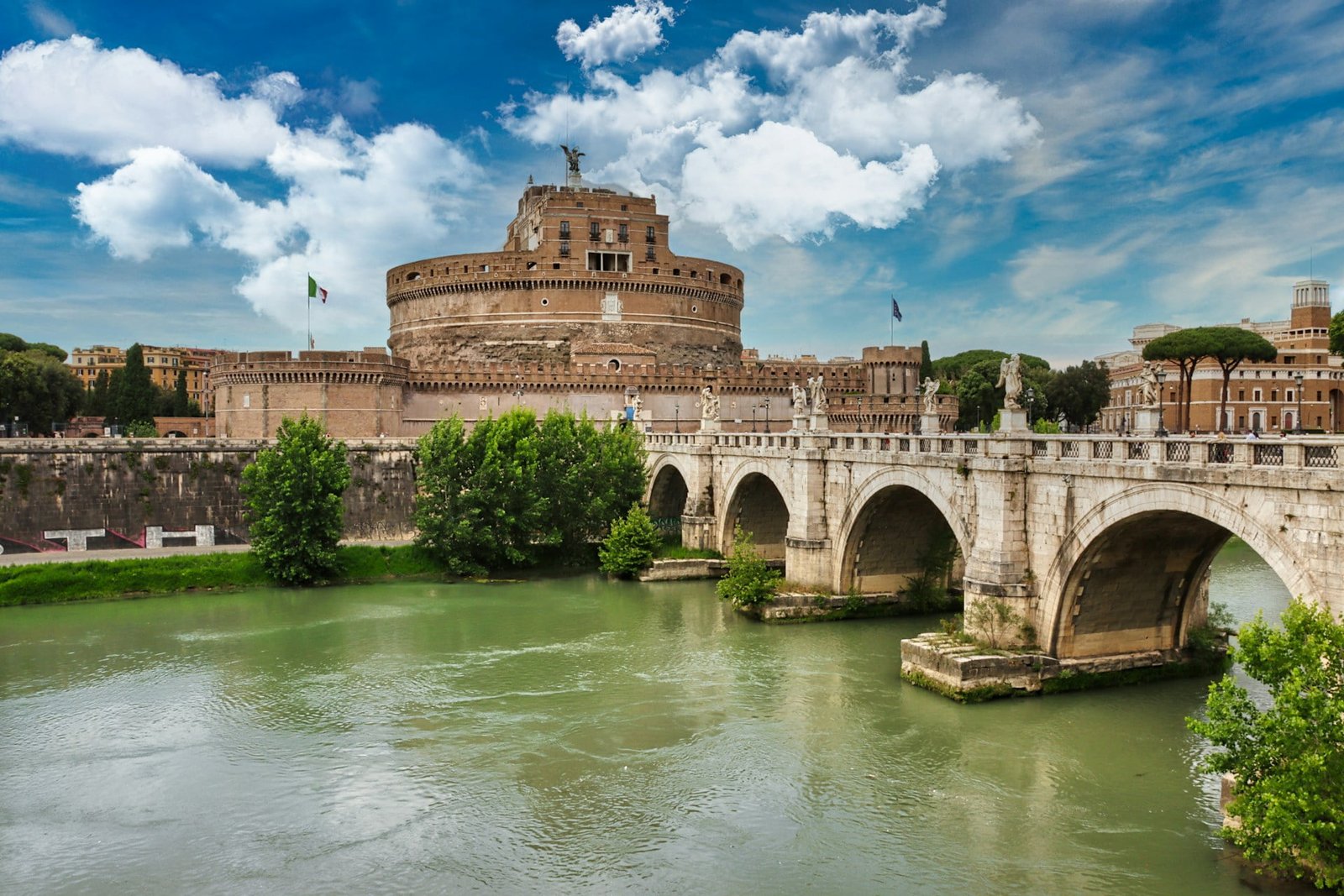 View of Castel Sant'Angelo in Rome, Italy, with the Ponte Sant'Angelo bridge spanning the Tiber River in the foreground, under a partly cloudy sky.