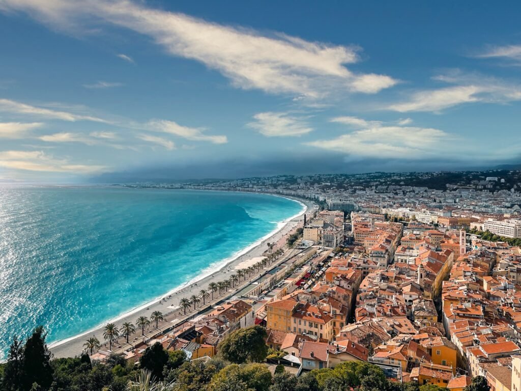 Aerial view of Nice, France, showcasing a coastal city with a long beach, palm trees, and numerous buildings under partly cloudy skies that epitomize the region's pleasant weather.