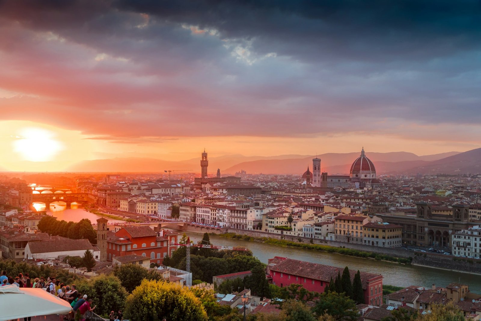 A sunset view of Florence, Italy, showing the Arno River, Ponte Vecchio, the Florence Cathedral with its prominent dome, and a crowd gathered in the foreground.