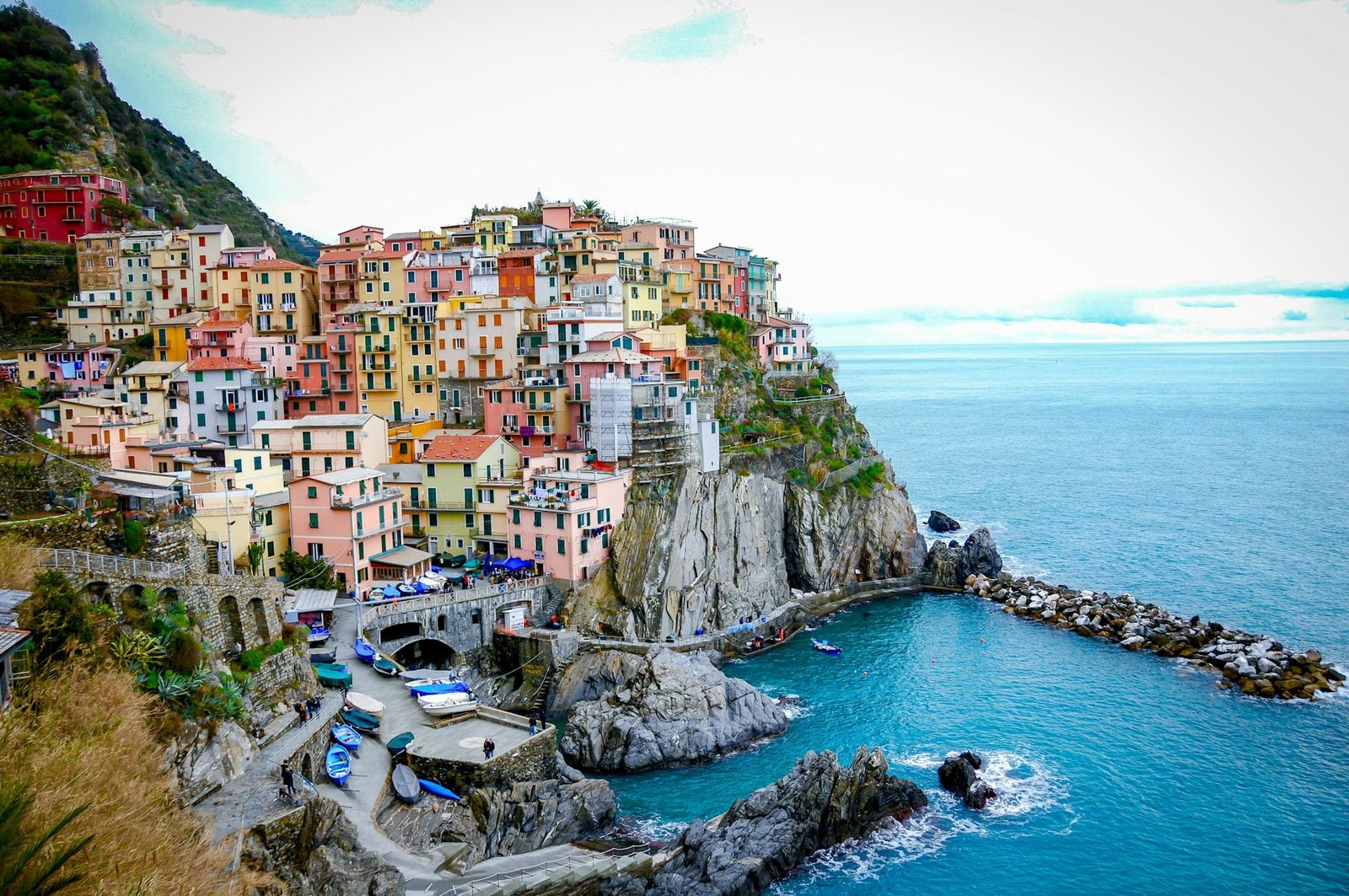 View of a coastal village with colorful buildings on a rocky cliff overlooking a blue sea, small boats docked in the water, and a road winding through the area. Experience this picturesque charm as part of the Best of Italy in 7 Days tour.