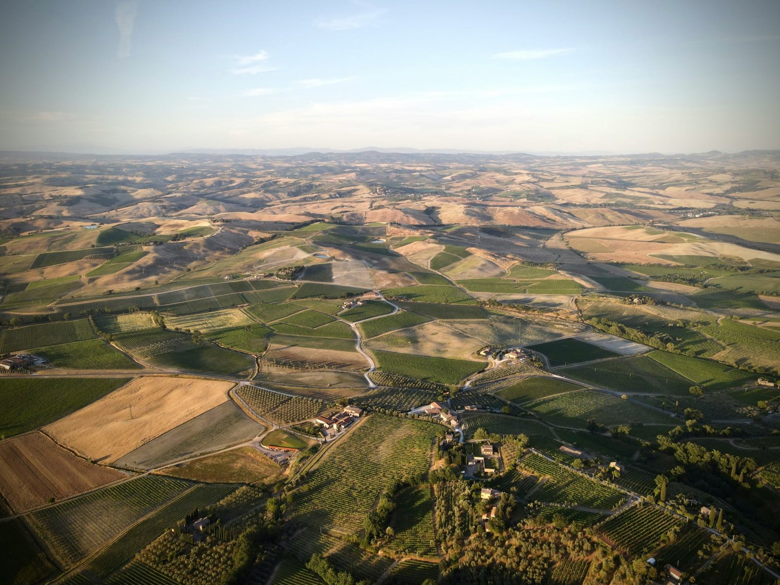 Aerial view of a vast, rolling countryside landscape with patchwork fields, small clusters of buildings, and winding roads under a clear sky—an idyllic scene for those exploring the Best of Italy in 7 days.