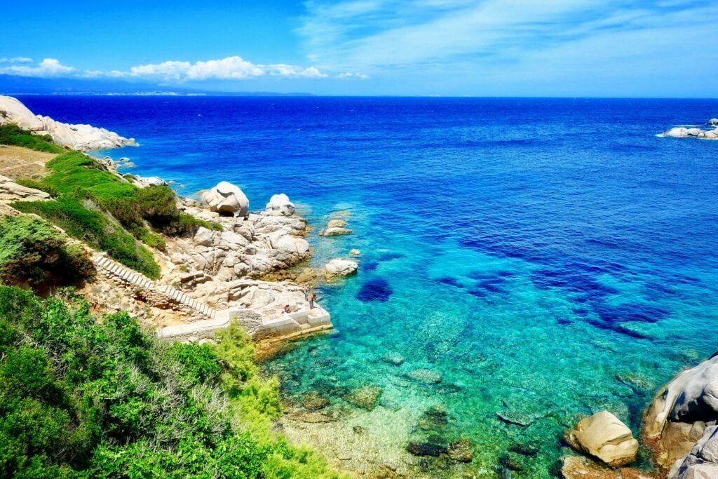 A rocky coastline with clear turquoise water, green shrubs, and a stone staircase leading down to the water. The bright blue sky is partly cloudy—one of the many things to do in Sardinia awaits you here.