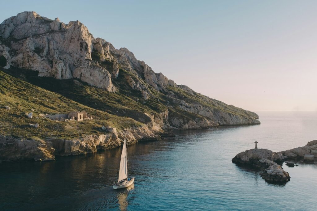 A sailboat glides through calm waters near a rocky coastline with a small stone structure, under the clear sky typical of weather in Marseille.