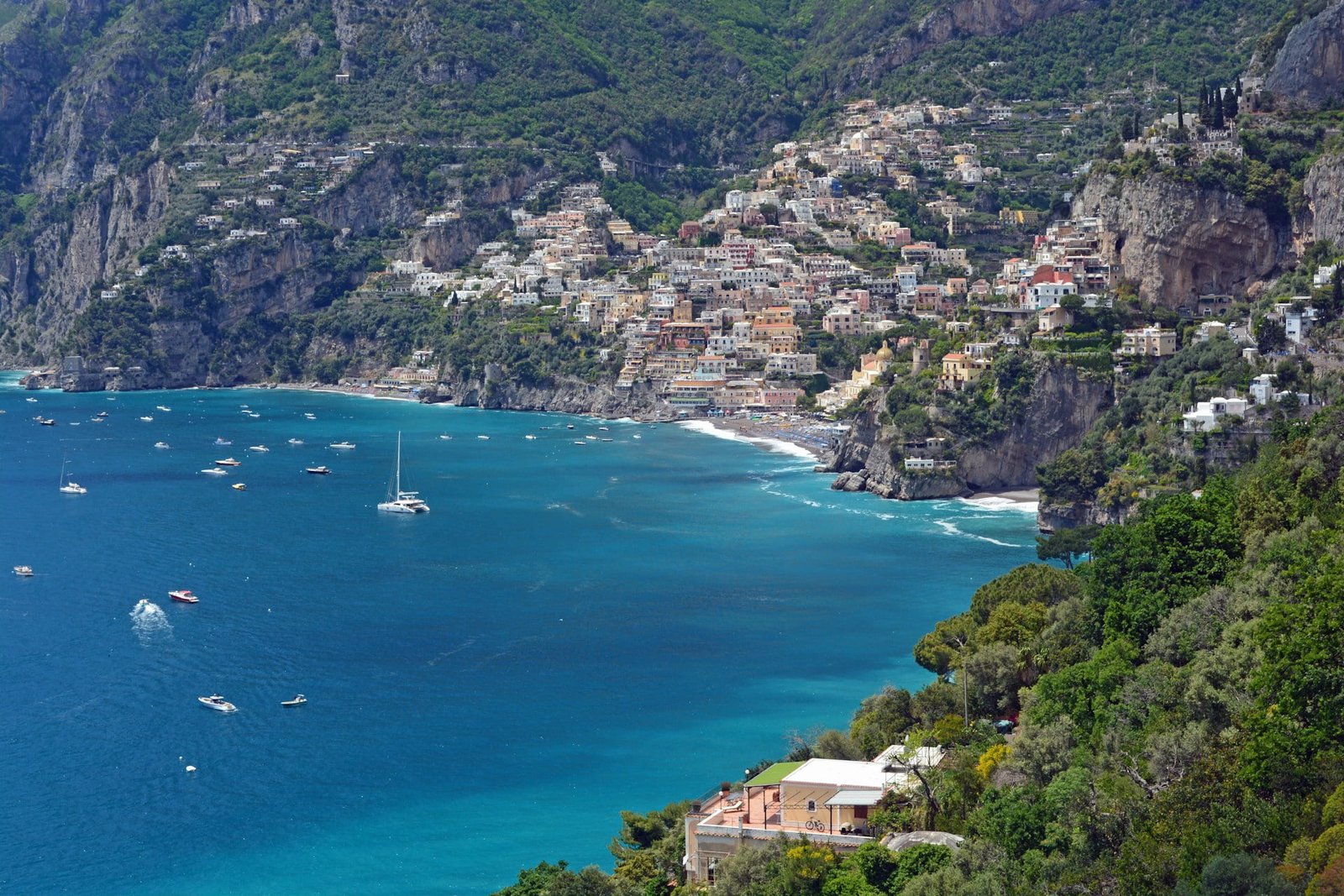 A cliffside town with numerous buildings overlooks a bay with several boats and yachts on deep blue water. Lush greenery surrounds the area.