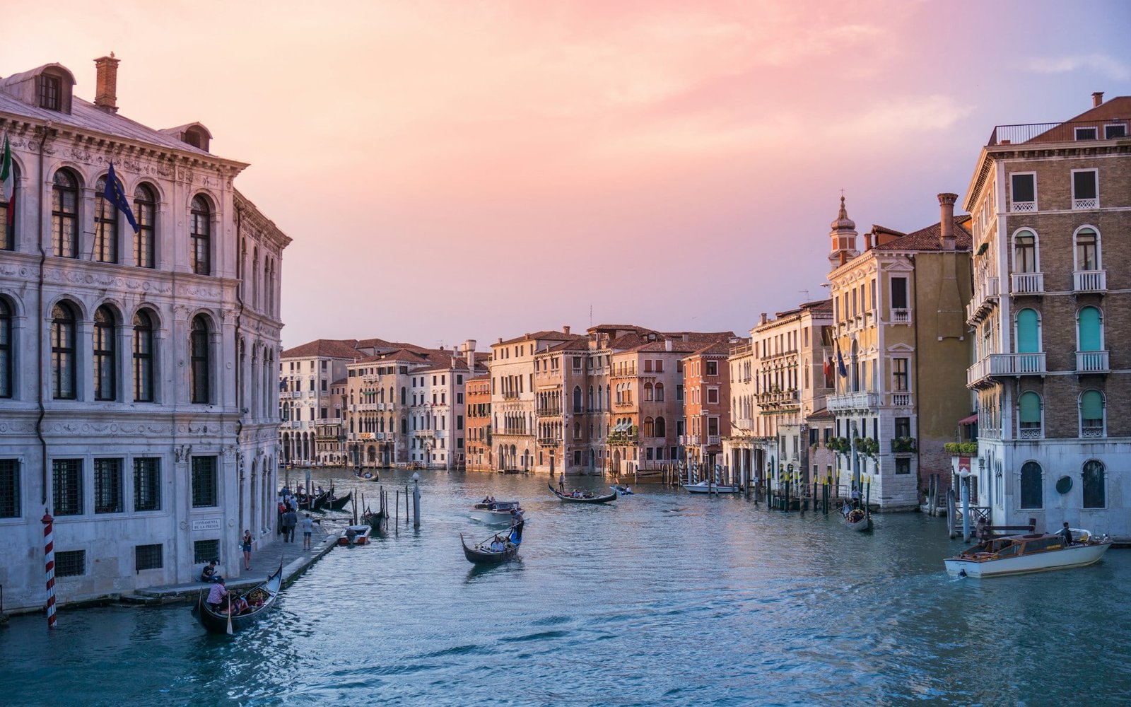 A canal in Venice with boats and gondolas floating on the water, surrounded by historic buildings and bathed in the soft light of a sunset.