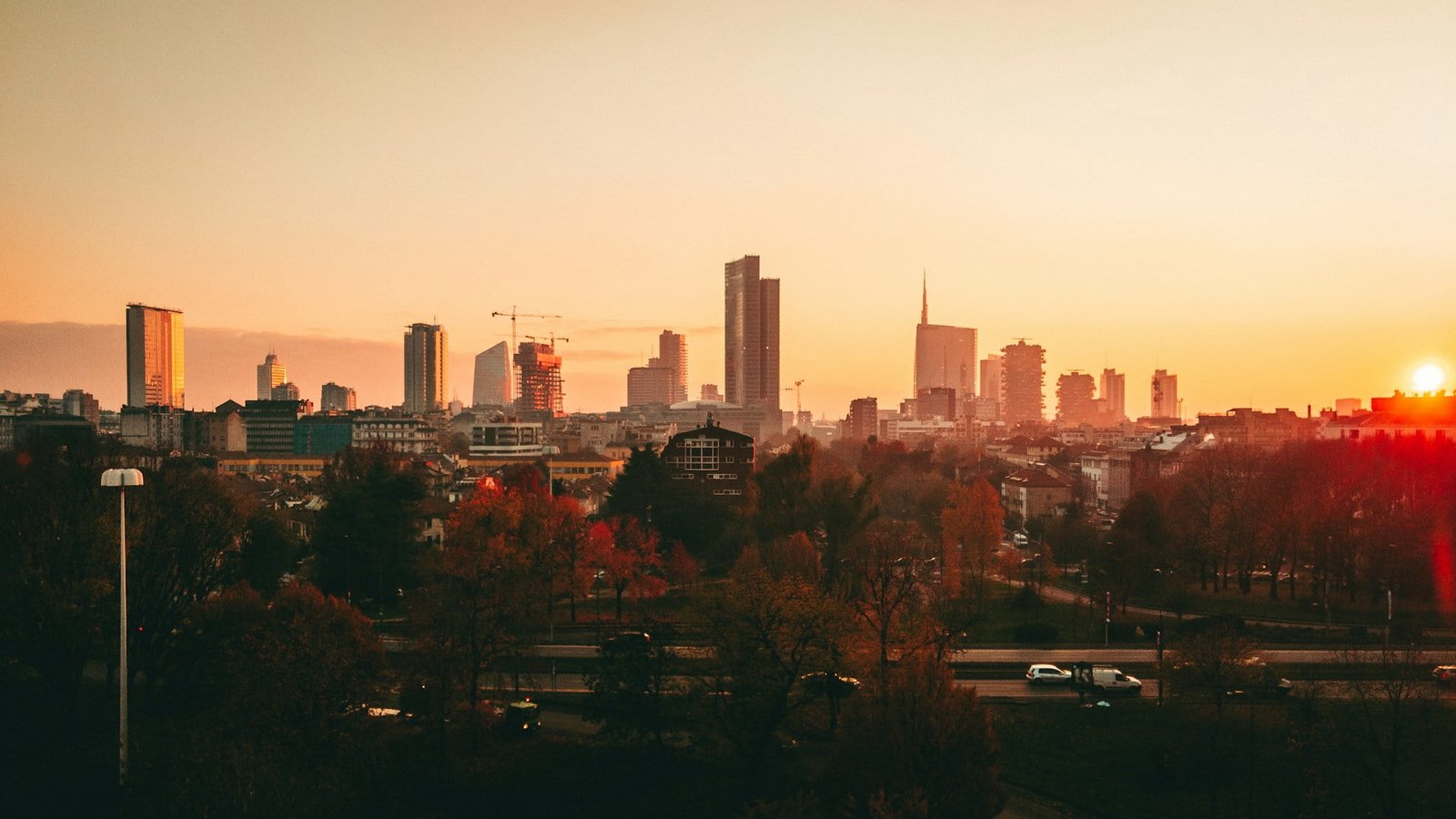 A cityscape at sunset with tall buildings and cranes in the distance. Below, a park with trees and a road with moving cars can be seen. The sky has an orange hue from the setting sun, reminiscent of the best time to visit Lake Como or explore Milan’s vibrant streets.