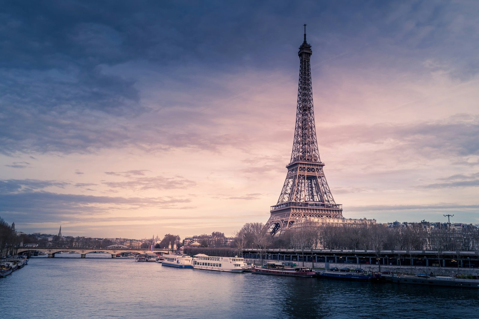 The Eiffel Tower stands tall under a cloudy sky at dusk, with the River Seine flowing in the foreground and several boats docked along the riverside, offering just one of many reasons to travel to France.