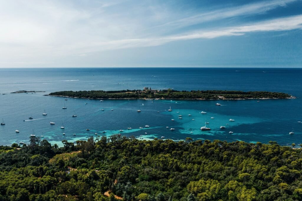Aerial view of a small island surrounded by clear blue waters and numerous boats, with a lush green forest in the foreground, reminiscent of a breathtaking European destination.