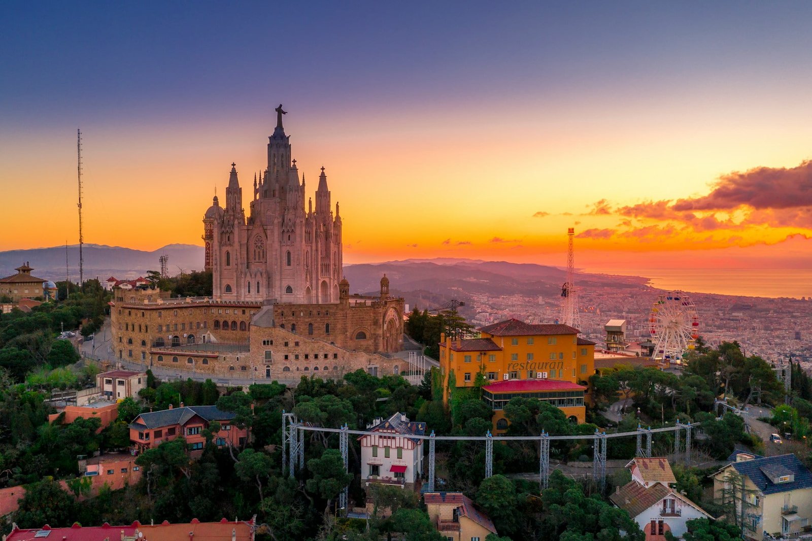 A large hilltop church overlooks a city at sunset, with various buildings and a Ferris wheel in the foreground, offering one of the best times to visit Barcelona.