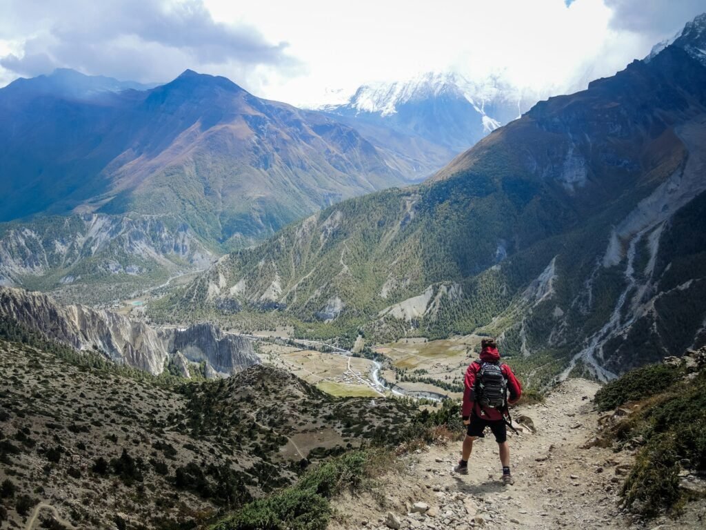 A person wearing a backpack and red jacket stands on a mountain path, overlooking a vast valley with green vegetation and rugged peaks under a cloudy sky.