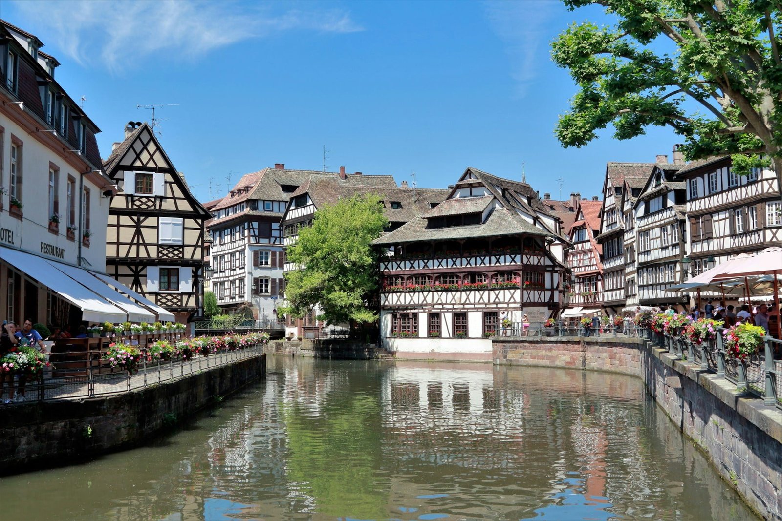 A picturesque canal surrounded by traditional half-timbered buildings and leafy trees under a clear blue sky.