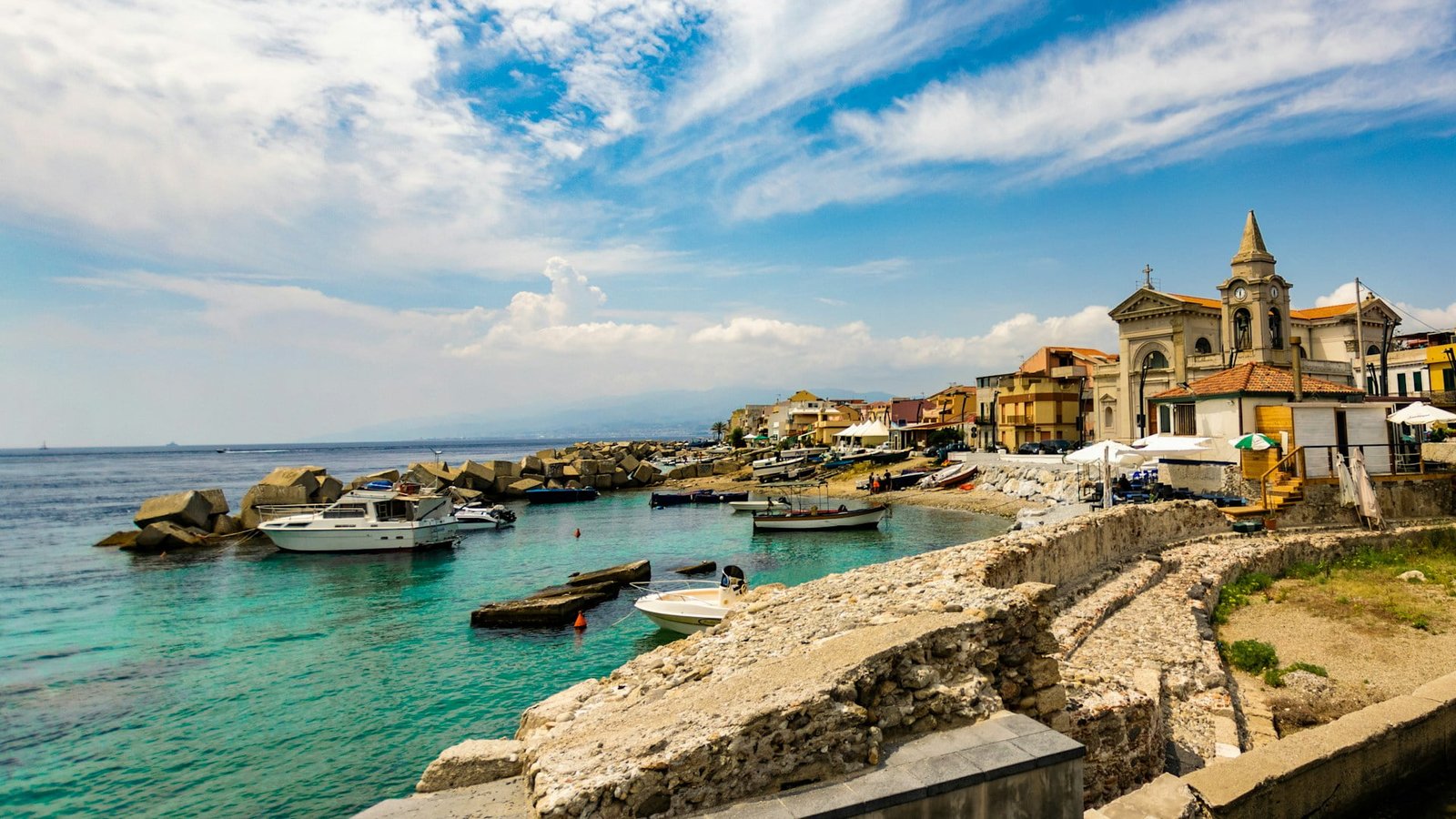 Coastal town with clear blue waters, boats docked near shore, historic buildings, and a church with a bell tower under a partly cloudy sky—truly a breathtaking European destination.