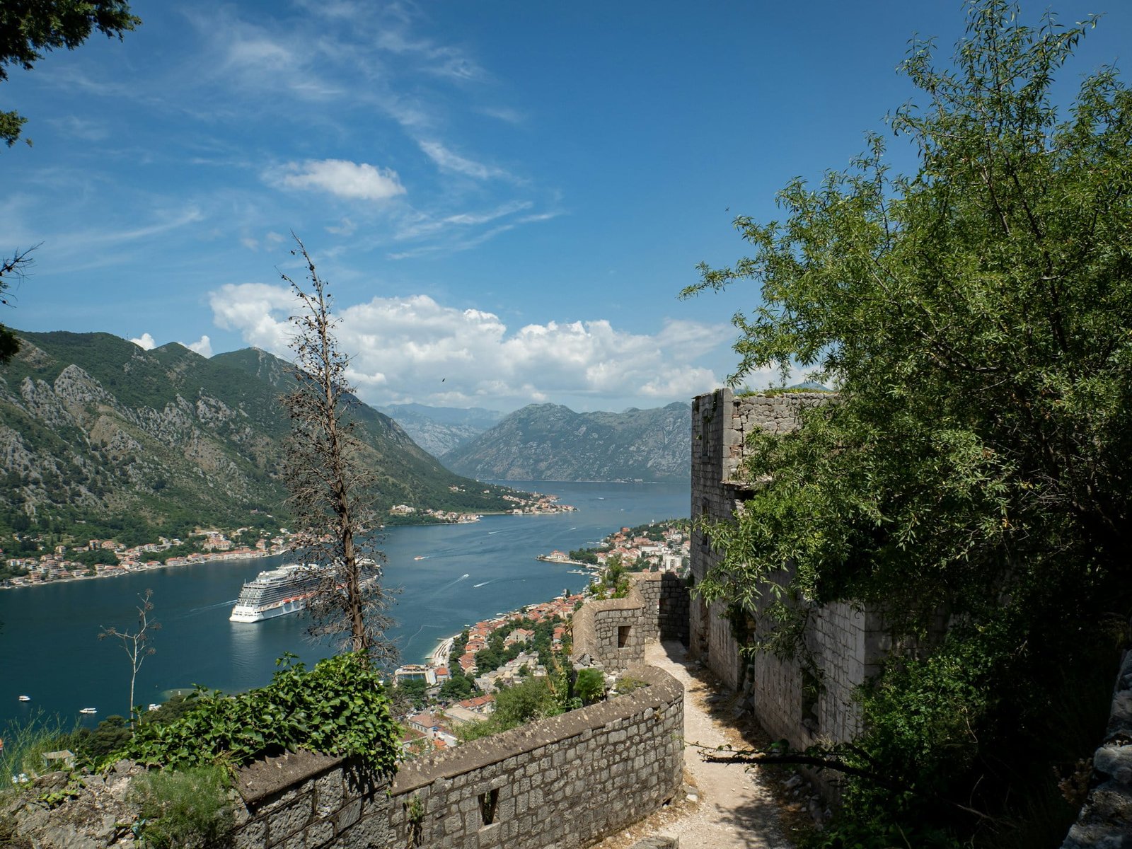 A stone pathway leads to a scenic view of a bay with a cruise ship, surrounded by mountains and an underrated European coastal town under a blue sky with scattered clouds.