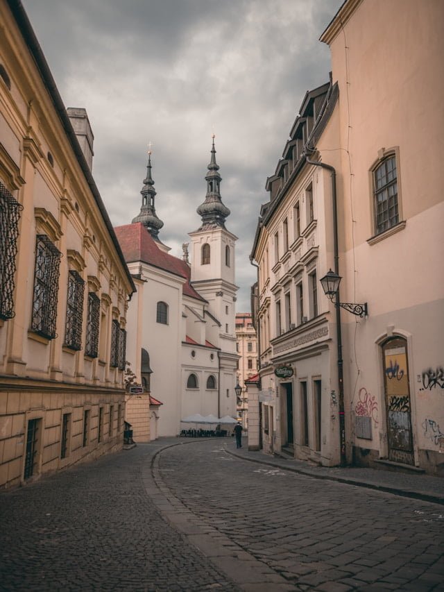 A narrow cobblestone street lined with historic buildings leads to a distant church with twin spires under a cloudy sky, capturing the charm of underrated European destinations.