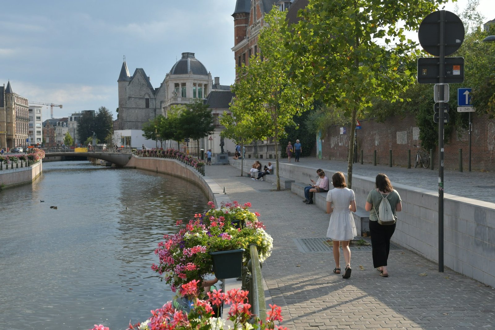 People walk along a canal lined with flowers in an underrated European destination. Historic buildings and a bridge are visible in the background, adding to the charm of this hidden gem.