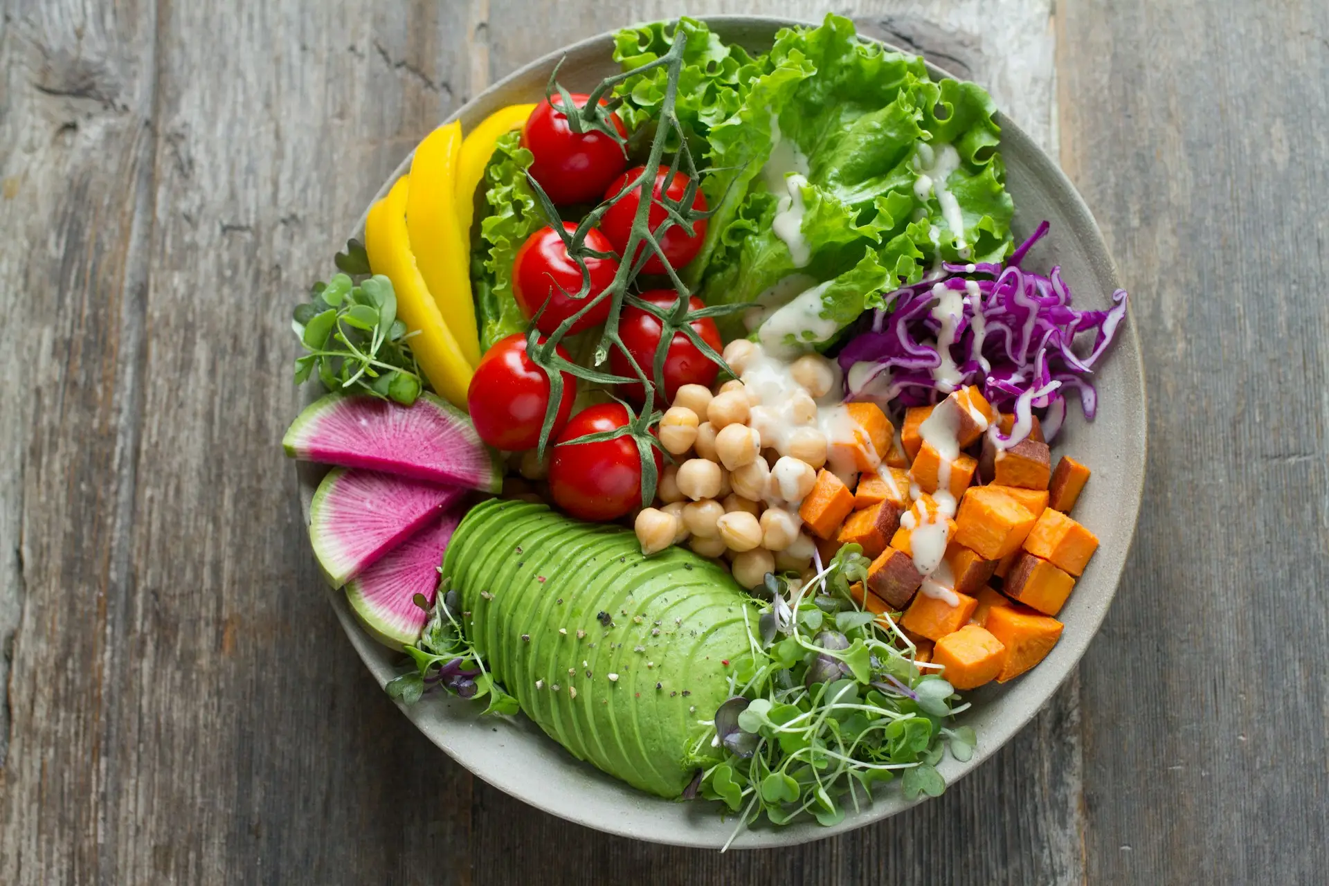 A vibrant and colorful vegan meal presented on a rustic wooden table, showcasing a variety of fresh vegetables and legumes. The dish includes ripe avocado slices, yellow bell pepper, cherry tomatoes, green lettuce, purple cabbage, watermelon radish slices, chickpeas, and diced sweet potatoes. This image exemplifies the diverse and appetizing options available for vegan travel in Europe.