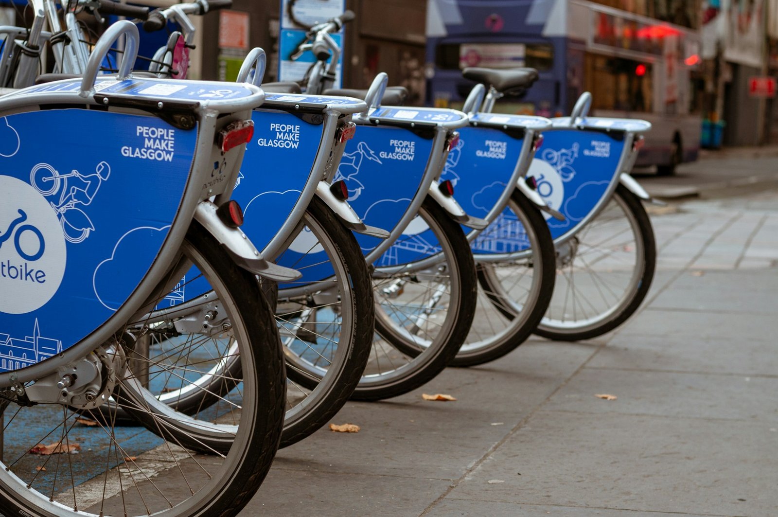 A row of blue bicycles from the ‘People Make Glasgow’ bike-sharing program, parked at a docking station on a city street, promoting sustainable travel in Europe. The bicycles are available for public use and encourage eco-friendly transportation, aligning with vegan travel values of minimizing environmental impact.