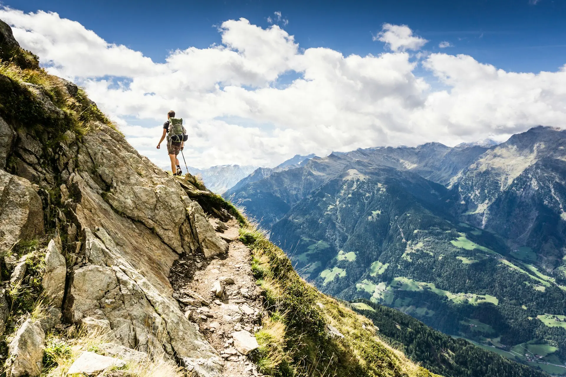 Being a Responsible Tourist, a hiker with a backpack walks along a narrow dirt trail on the edge of a mountain. The trail overlooks a scenic view of lush green valleys and rugged mountain ranges under a partly cloudy sky, emphasizing the importance of preserving such natural beauty while exploring.