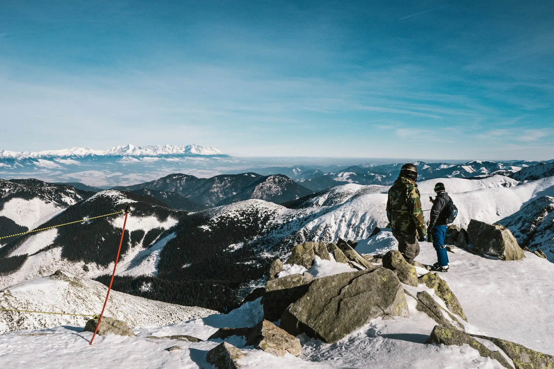 winter sports destinations in Europe Two people stand on a snowy mountain, looking at other snow-covered mountains and hills under a clear blue sky. A red and yellow ski marker shows this is a popular spot for winter sports destinations in Europe.