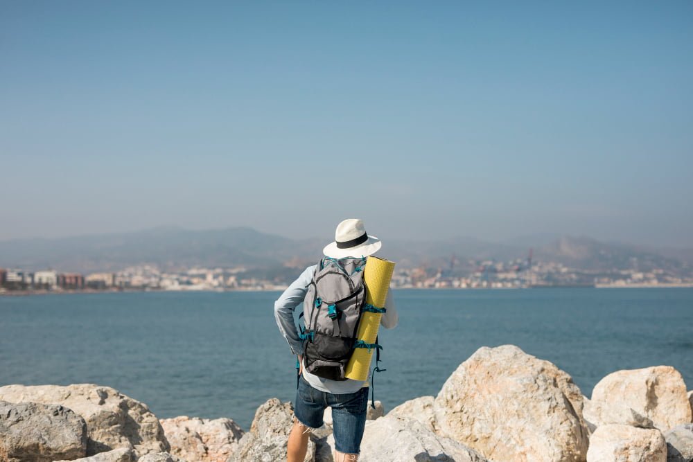 Back view of a traveler with a backpack and rolled-up map standing on rocky terrain, gazing towards a coastal European cityscape under a clear blue sky, symbolizing the start of a Multi-City Trip to Europe.