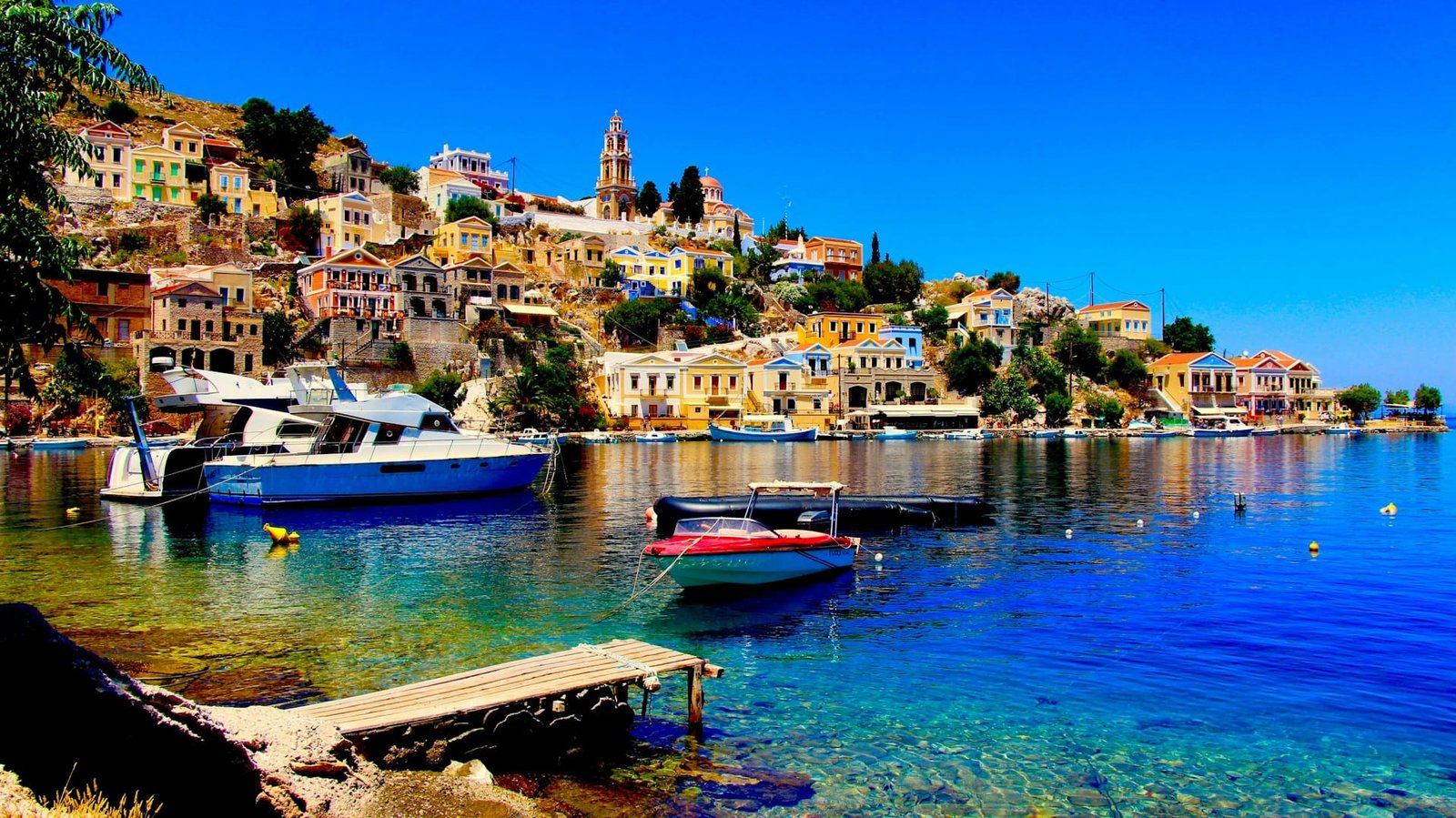 Visiting Greece for the first time and seeing colorful houses on a hillside in Halki, with boats in the clear blue water and a bright sky.