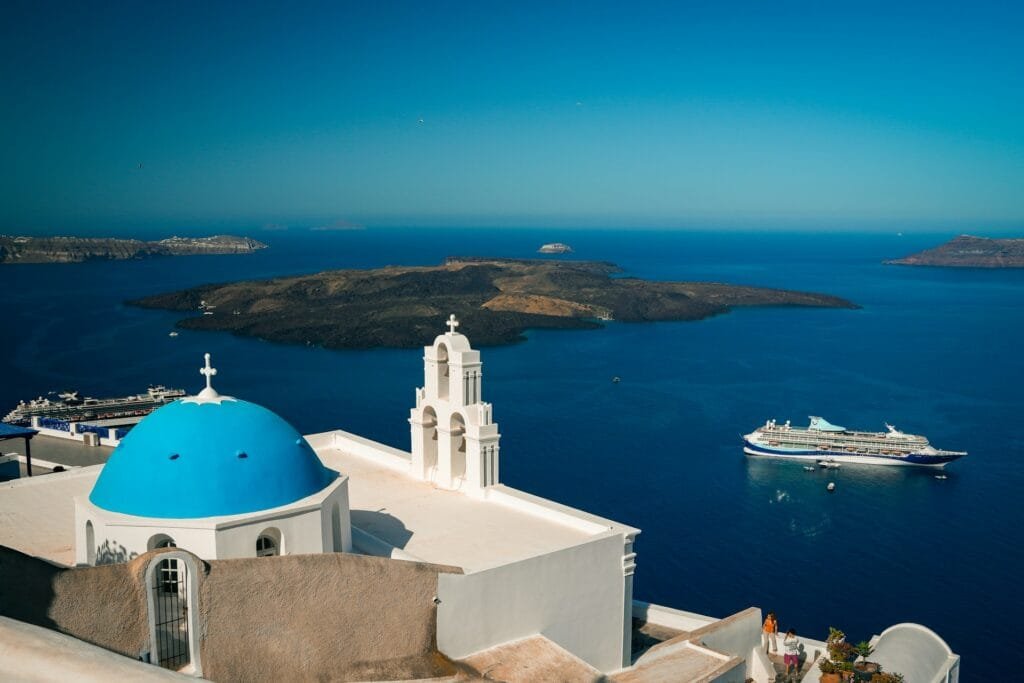 A breathtaking view of Santorini, Greece, showcasing the iconic blue dome of a church against a backdrop of the deep blue Aegean Sea with a cruise ship and smaller boats nearby, under a clear sky. Perfect for those visiting Greece for the first time.