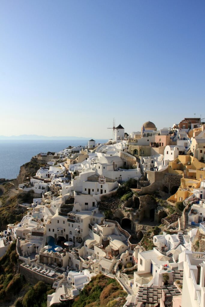 Explore Greece: A scenic view of the iconic white-washed buildings with blue domes in Santorini, Greece, cascading down the cliffside towards the Aegean Sea, with a traditional windmill standing prominently against a clear sky.