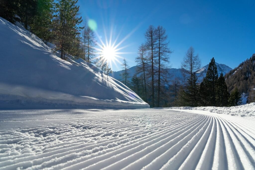 A pristine snow-covered landscape with a clear blue sky, the sun shining brightly, and well-defined ski tracks leading through the trees, suggesting it’s not the worst time to visit Switzerland for winter sports enthusiasts.