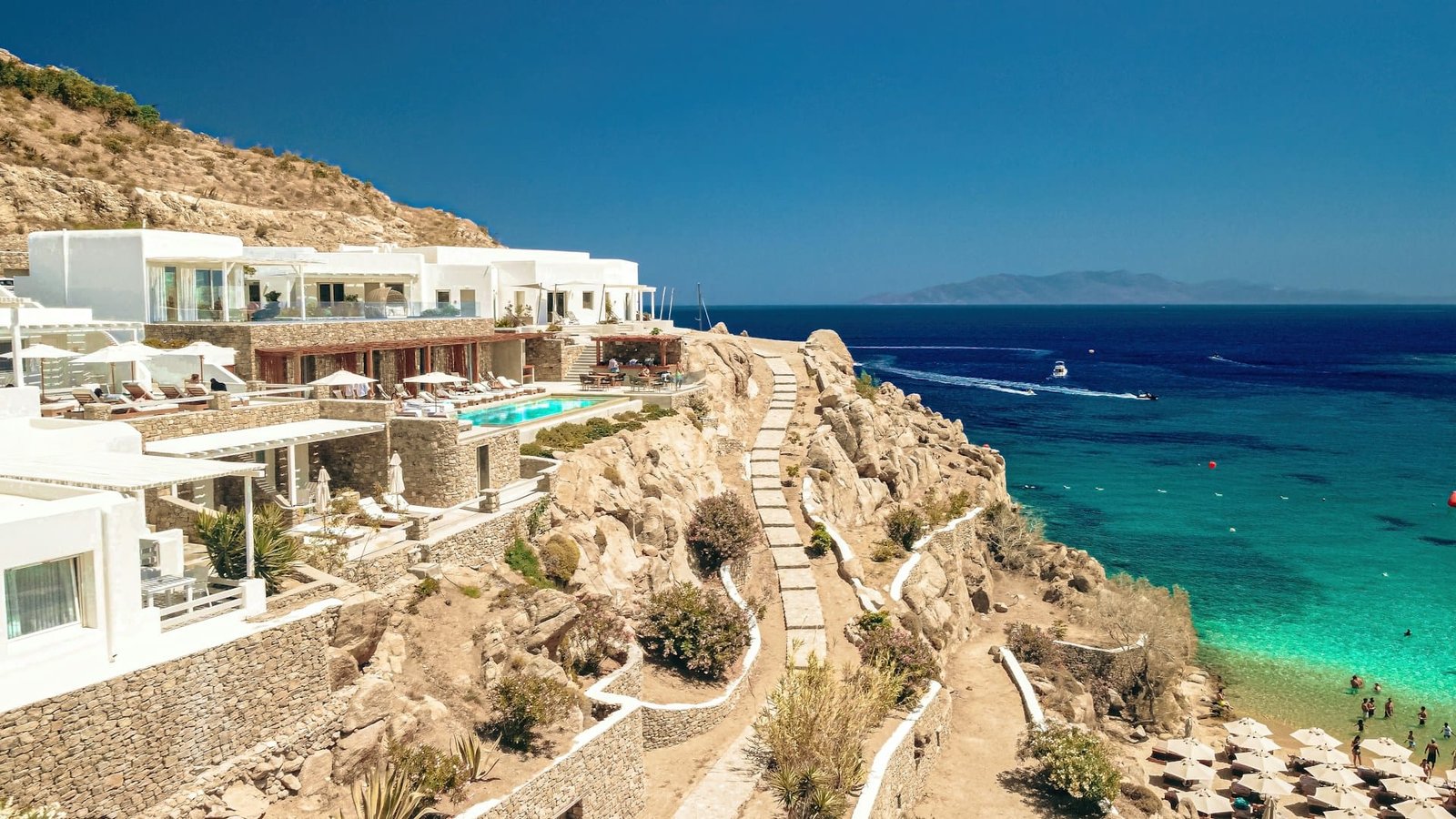 A panoramic view of Mykonos, Greece, showcasing the iconic white-washed architecture with blue accents perched on rocky cliffs overlooking the crystal-clear Aegean Sea. A stone pathway meanders along the cliffside, leading to various terraces and balconies that offer breathtaking views, embodying the quintessential experience of visiting Greece for the first time.