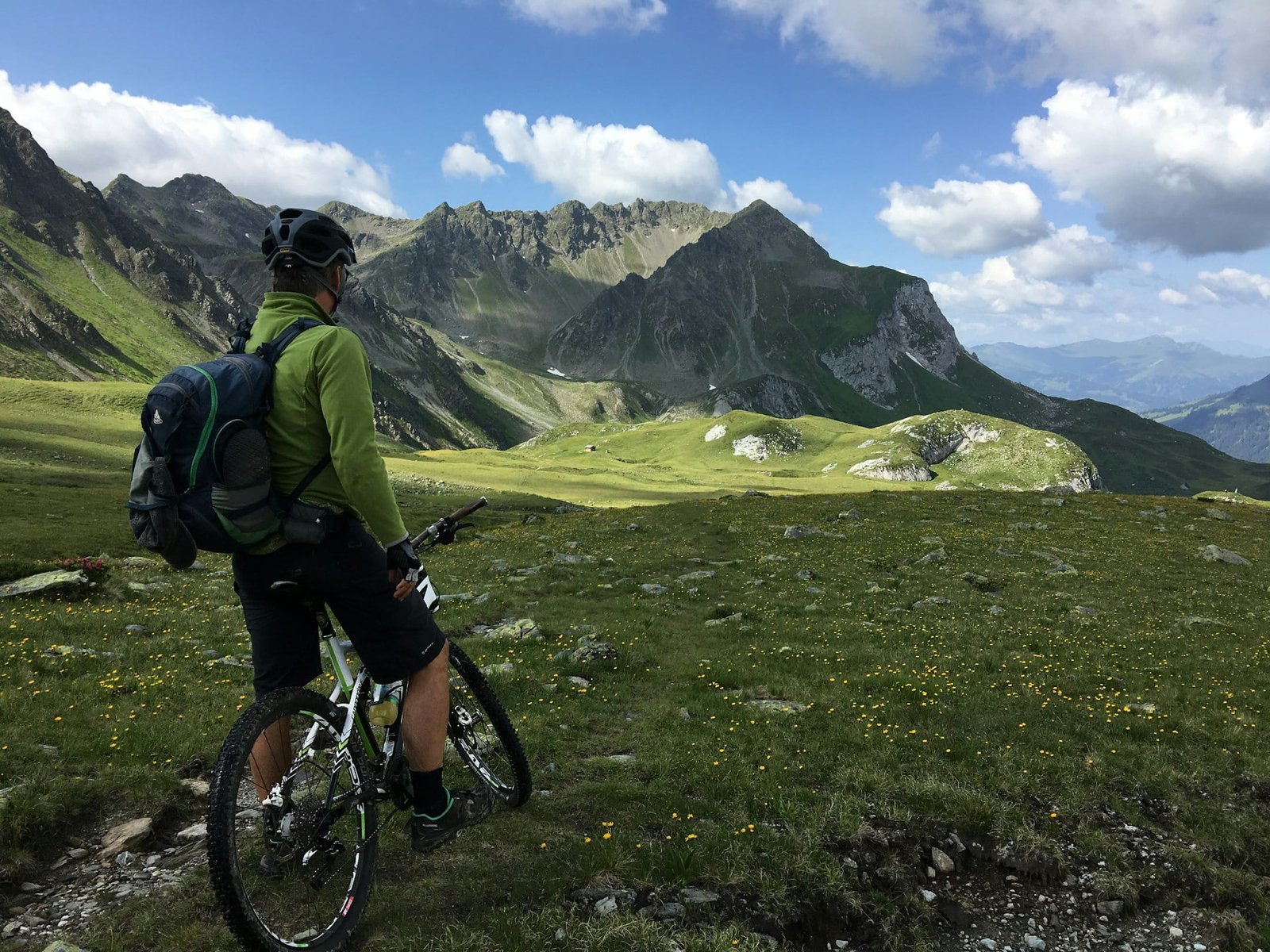 A cyclist in a green jacket and helmet stands with a mountain bike, gazing at the majestic Swiss Alps under a clear blue sky, surrounded by lush green meadows dotted with yellow wildflowers. Plan a Trip to Switzerland for this breathtaking view.