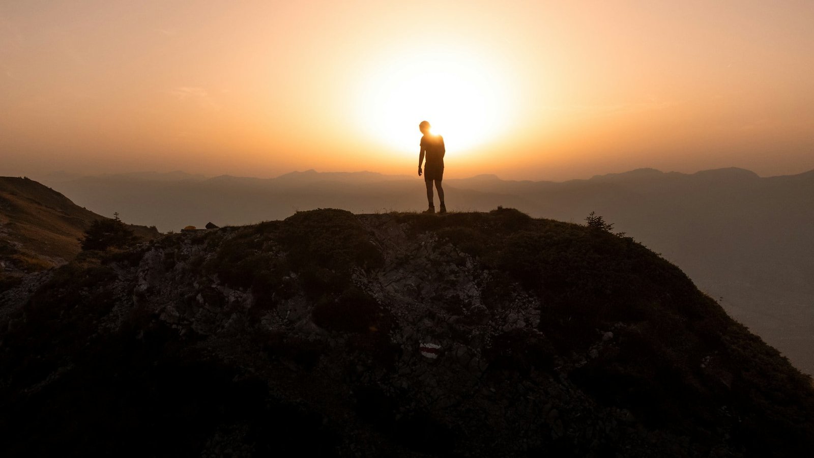 Plan a trip to Switzerland and witness a silhouette of a person standing on a hilltop in Arvigrat, Dallenwil, with the sun setting behind them, casting a warm glow over the hazy mountain ranges in the distance.