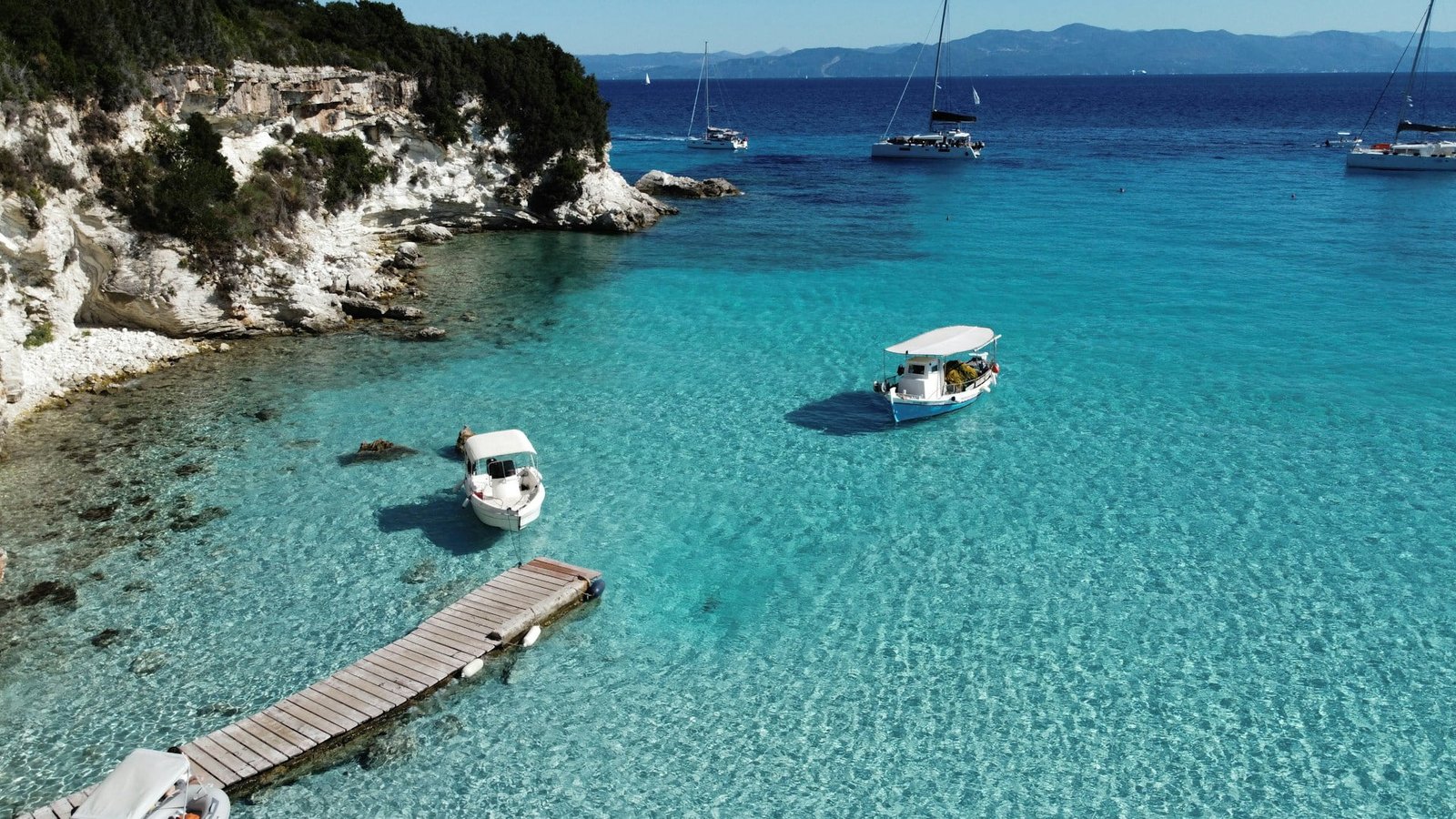 A serene view of Voutoumi Beach in Antipaxos, Greece, showcasing crystal-clear turquoise waters with boats anchored nearby and a wooden pier extending into the sea. The image contrasts the idea of the “worst time to visit Greece” with its inviting scenery.