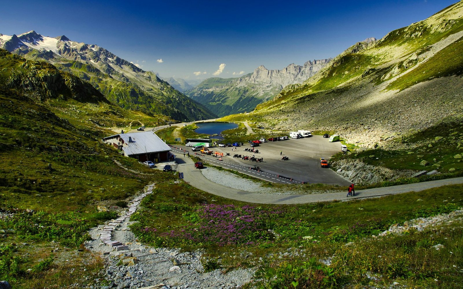 A scenic view of Sustenpass in Gadmen, Switzerland, showcasing a winding road leading to a mountain pass with a small building and parked cars. Lush greenery and rugged mountains under a clear blue sky make it a perfect starting point for those wondering where to go in Switzerland for the first time.