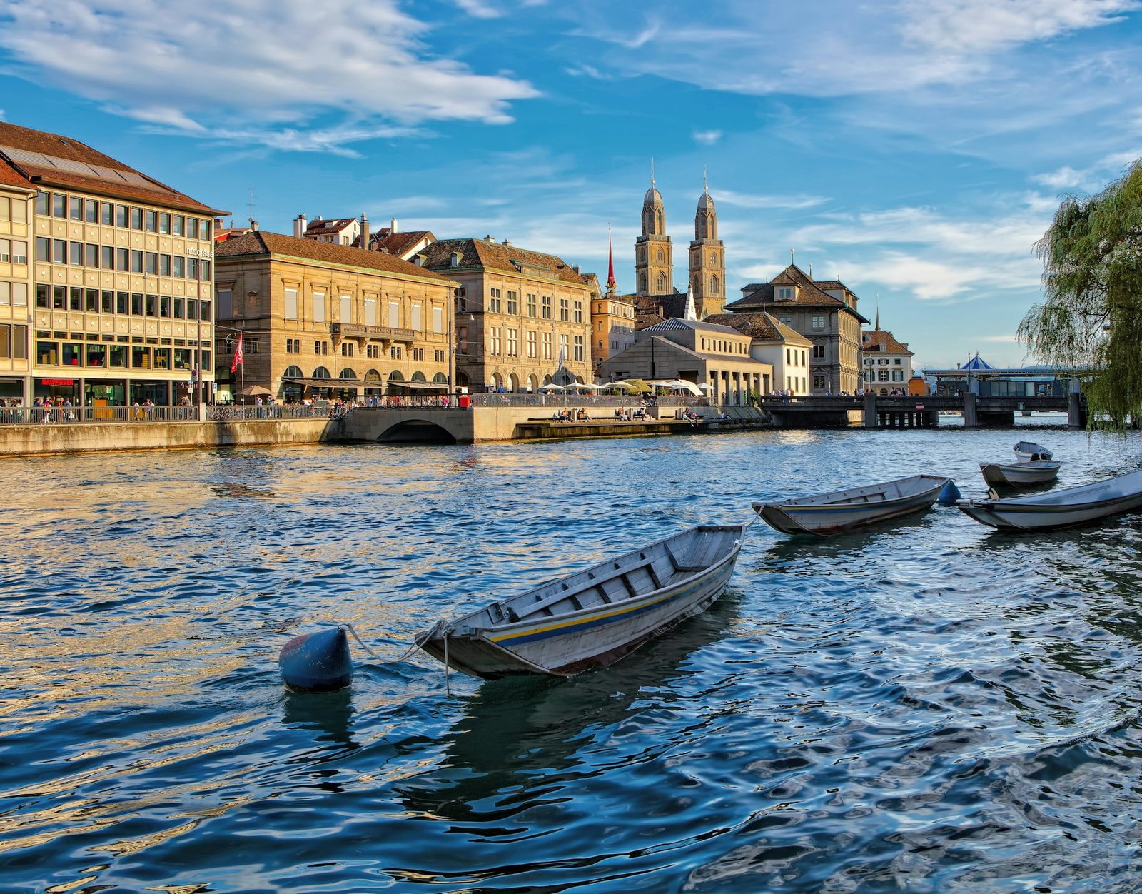 Plan a Trip to Switzerland: View of Zürich featuring the Limmat River with boats, historic architecture including the Grossmünster church, and a vibrant blue sky with wispy clouds.