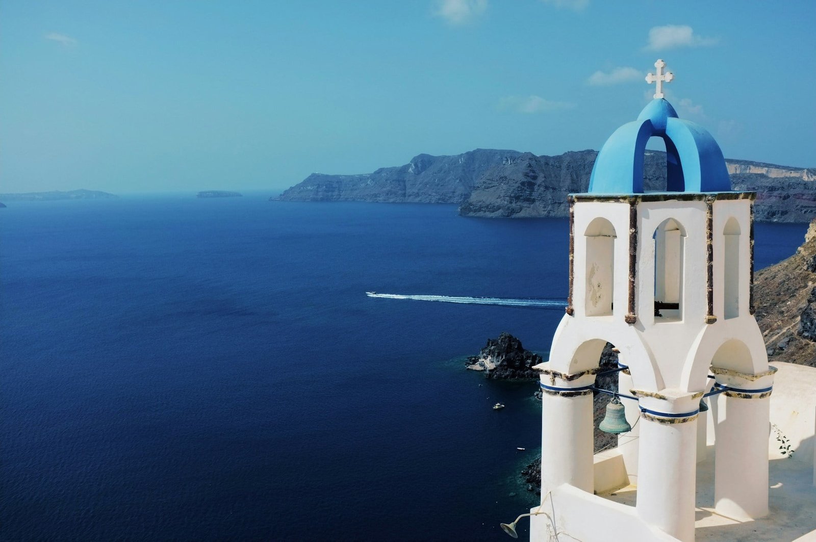 A stunning view of a traditional white and blue-domed church overlooking the deep blue waters of the Aegean Sea, with a distant rocky coastline under a clear sky, capturing the essence of exploring Greece's beautiful islands