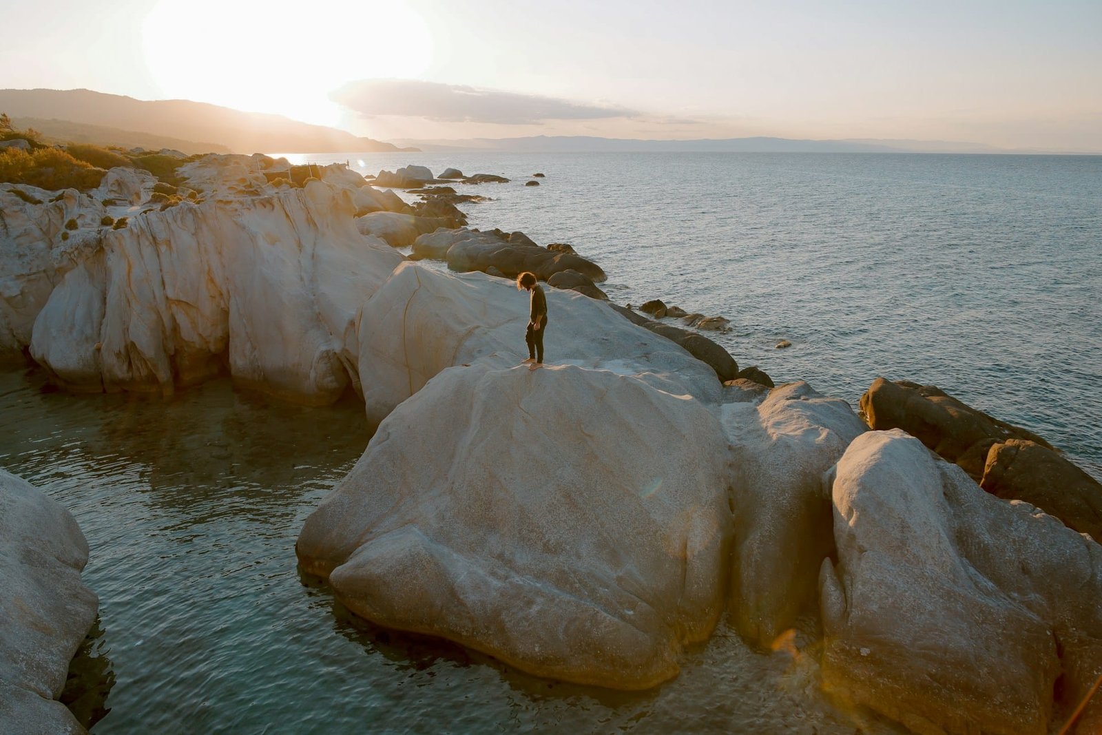 A breathtaking view of Orange Beach in Sarti, Greece, captured at sunset with warm golden hues bathing the rocky shoreline and a solitary figure standing atop a boulder, encapsulating the serene beauty one might encounter when visiting Greece for the first time.