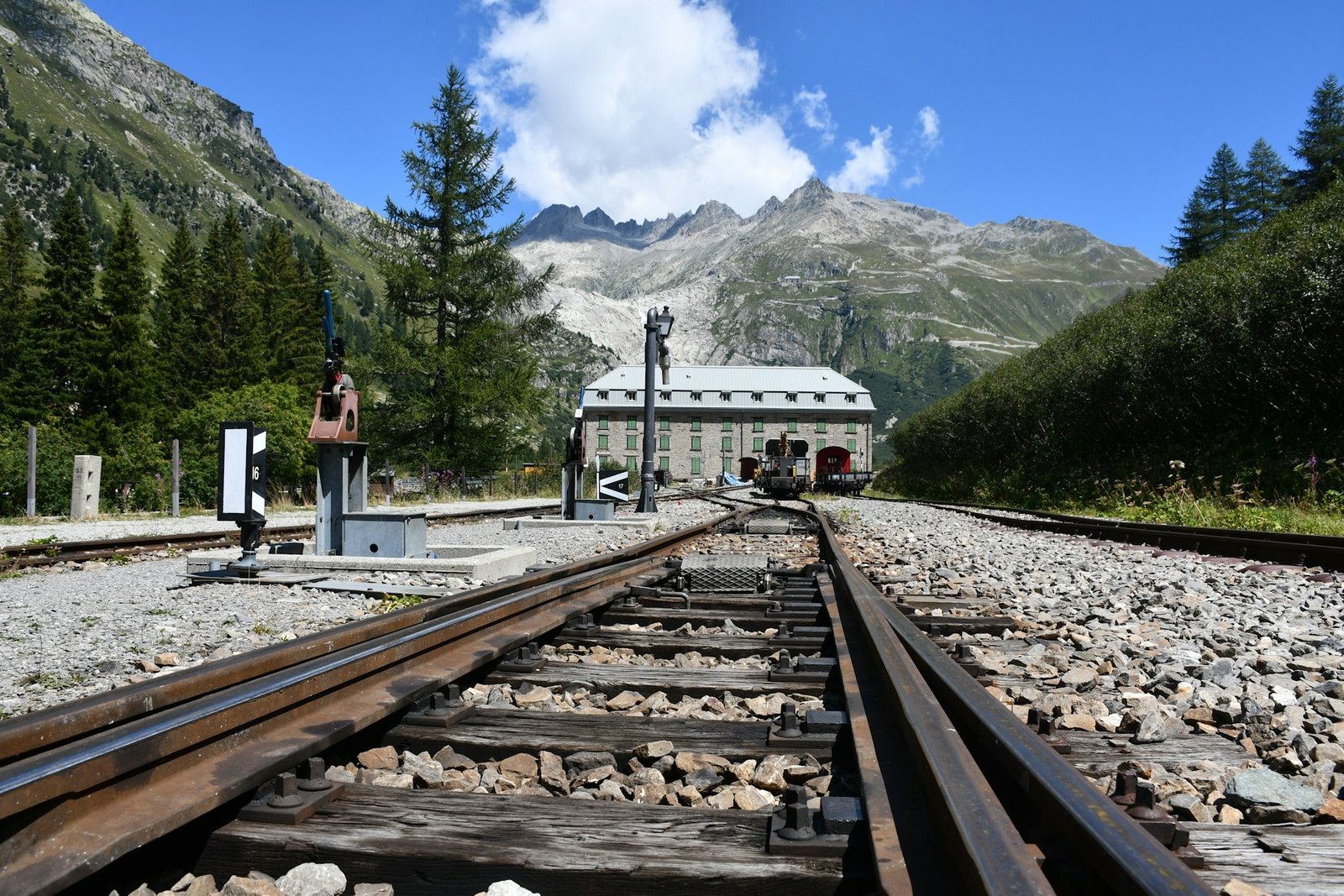 A scenic railway station in Gletsch, Obergoms, Switzerland, surrounded by green trees, rocky mountains, and a clear blue sky, showcasing what Switzerland is famous for—its breathtaking landscapes and mountain railways.