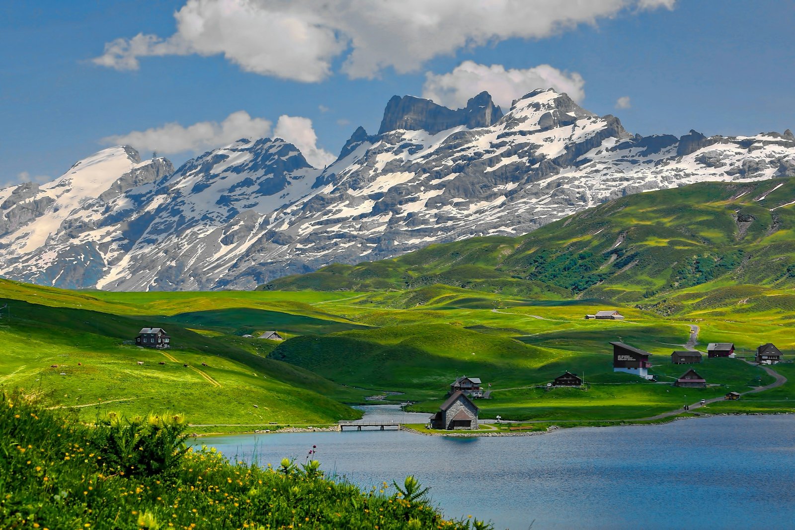 Scenic view of Melchsee-Frutt, Kerns, Switzerland with lush green fields, a tranquil lake, and towering snow-capped mountains in the background – showcasing one of the many reasons to visit Switzerland.