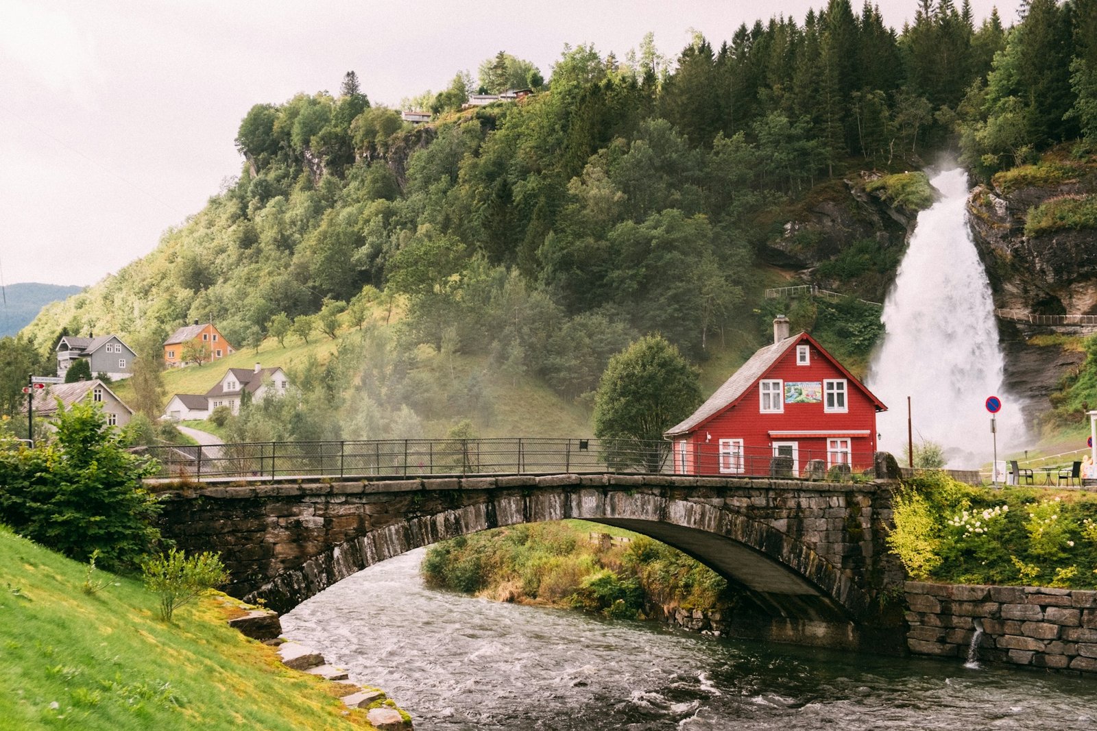 A scenic view of Steinsdalsfossen waterfall in Norheimsund, Norway, surrounded by lush greenery, quaint houses, and a stone bridge. Is Norway a good place to visit?