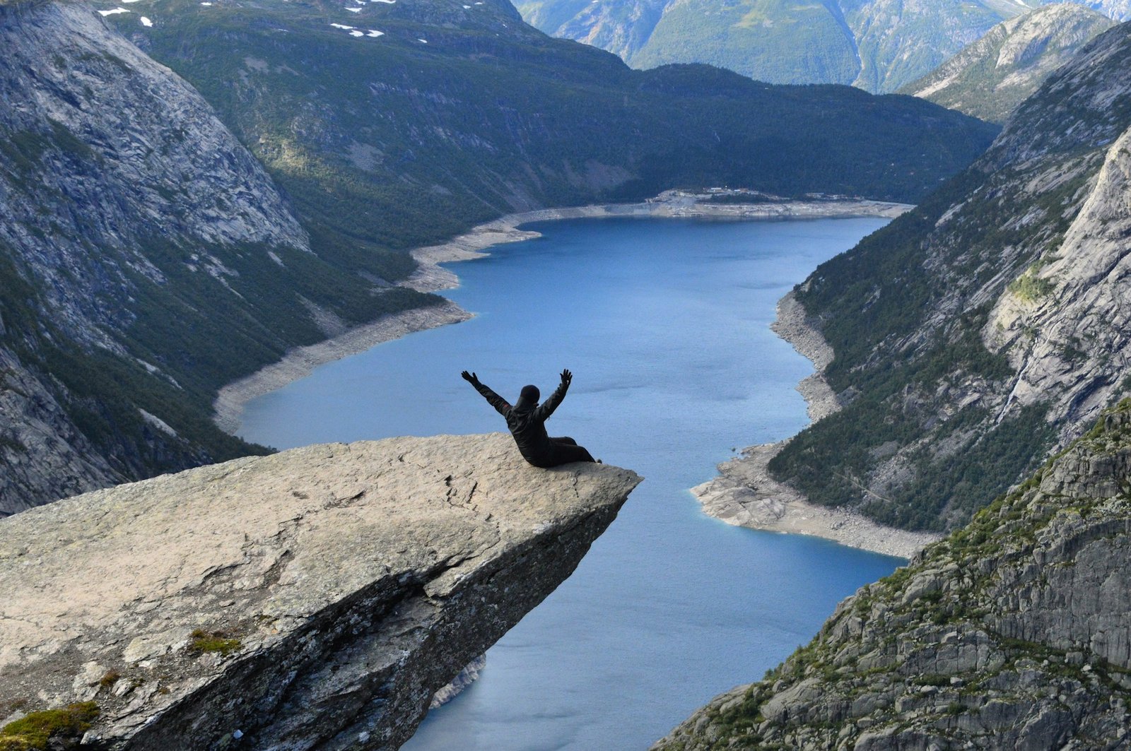 A person sitting on Trolltunga cliff with arms raised, overlooking a stunning blue fjord. Is Norway worth visiting? Absolutely!